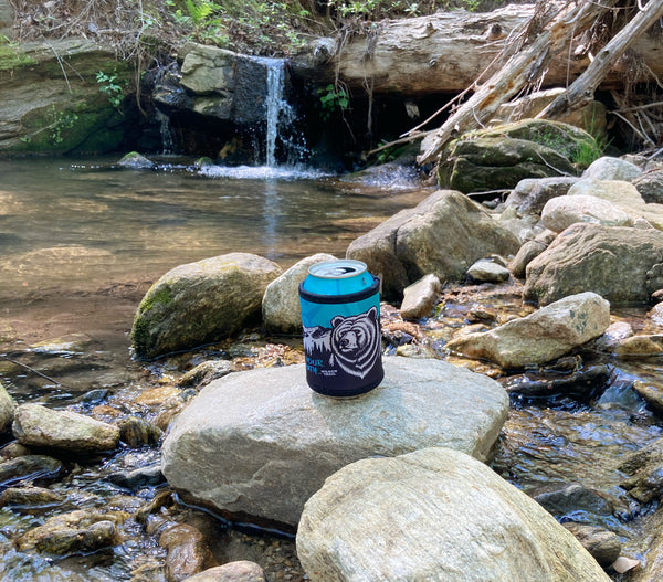 A blue and black can coozie with a white design sitting on a rock near a small waterfall.