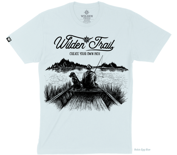 Light blue t-shirt with black graphic design of a man and lab fishing in a boat on a lake with mountains in the background. 'Wilden Trail Create Your Own Path text at the top of the design.