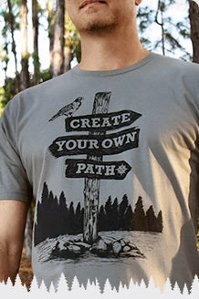 The image shows a man wearing a T shirt featuring a hawk perched on a directional wooden sign that reads “Create Your Own Path,” with rocks at the base of the post.